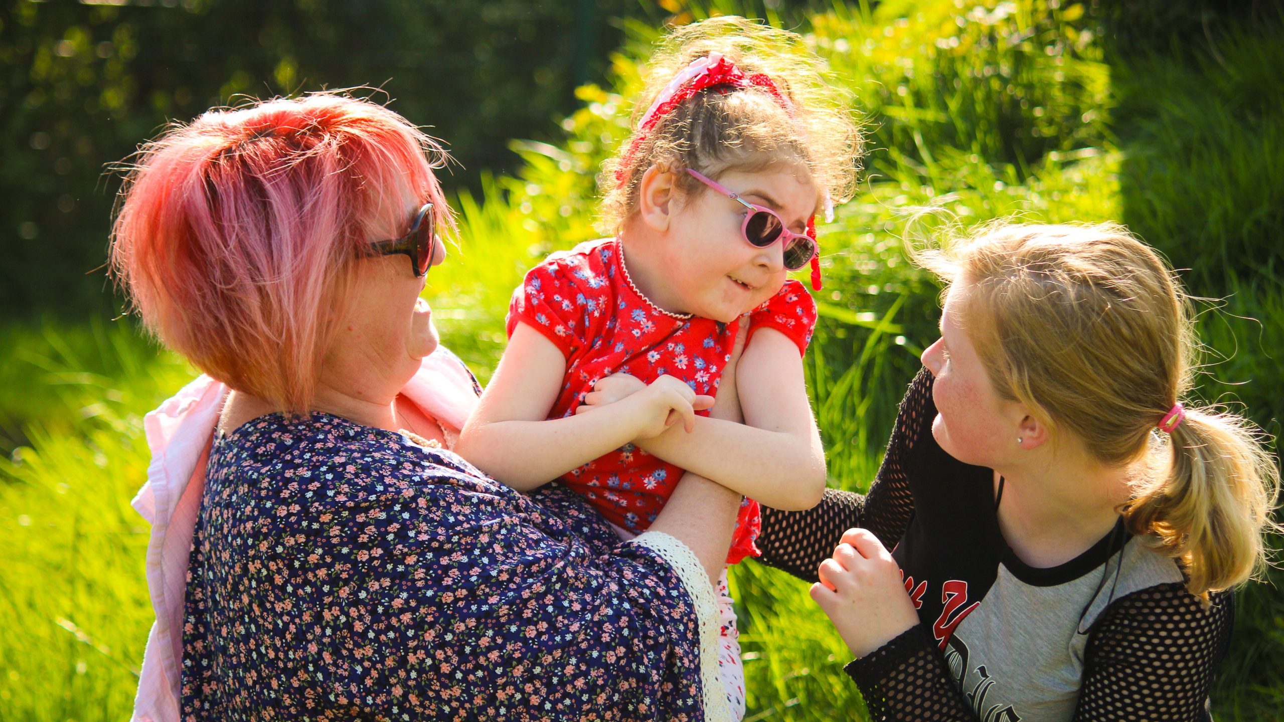 A family playing with each other. A mum is holding a child while another, slightly older, child stand beside playing with the girl in her mums arms. They all look amongst each other on a bright sunny day.