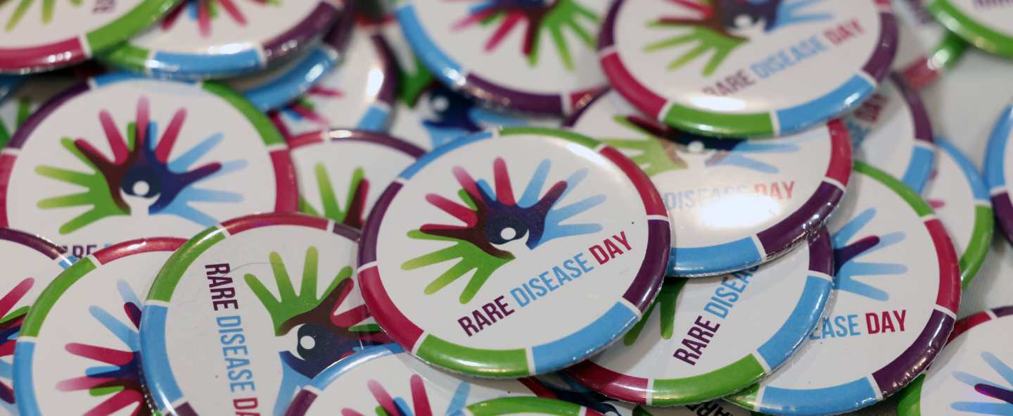 A pile of round badges for Rare Disease Day. The Rare Disease Logo is in the centre with the branded colours in segmented around the edge.