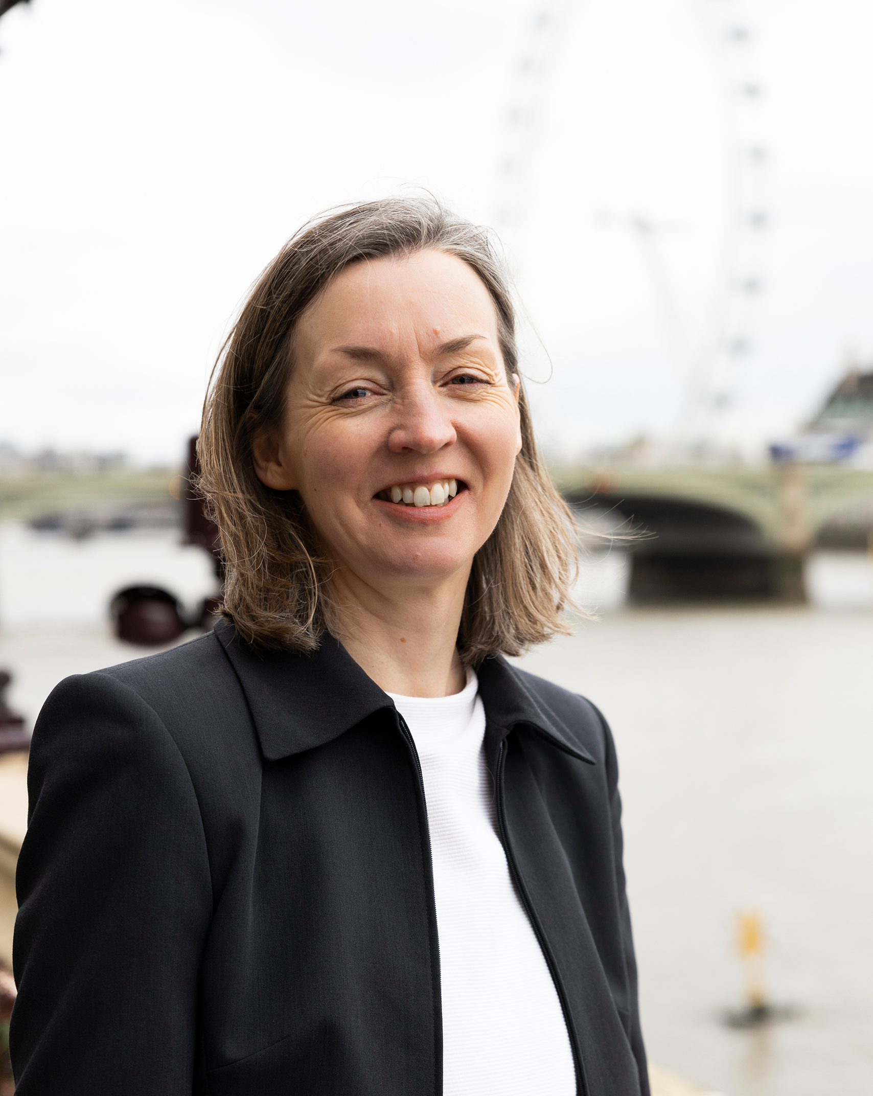 A headshot of Amy, from Genetic Alliance UK's Research team. She has shoulder length hair and smiles with a friendly smile. She stands by the Thames river at the Westminster Parliament building.