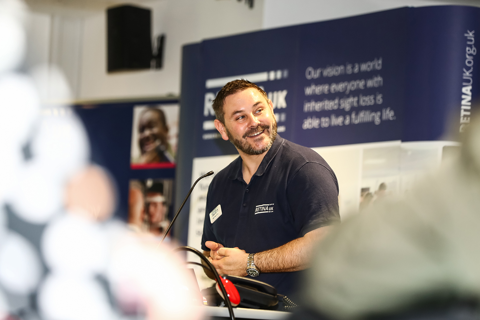 Matthew Carr, Strategic Lead – Campaigning, Influencing and Policy at Retina UK. He is speaking at an event standing in front of a small microphone. He looks over the side and smile. Behind you can see Retina UK boards and information, he also wears a navy blue Retina UK polo shirt.