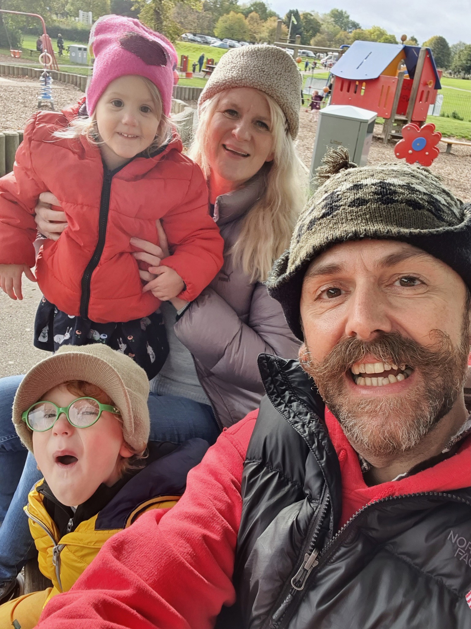 Stephen White, Jo Milne and family. They are all outside, Jo holds on child balanaved on her knee while crouching, Another young boy pulls a funny face wearing blue glasses. Steve takes the photo as a selfie. They all wear big warm coats as if it is a cold day.