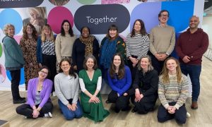 The Genetic Alliance UK team stood in front of a colourful background. a note on the wall, written in white in a black bubble, reads 'Together'