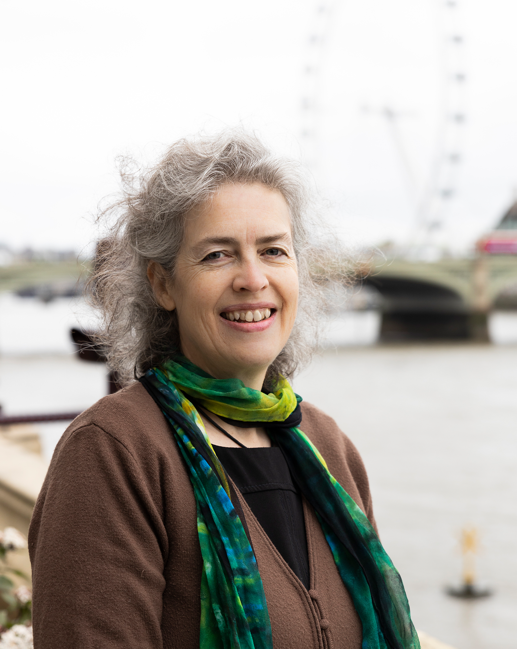A headshot of Jen, from Genetic Alliance UK's Research team. She has grey hair and smiles with a big friendly smile that shows her teeth. She stands by the Thames river at the Westminster Parliament building and wears a green scarf and brown cardigan.