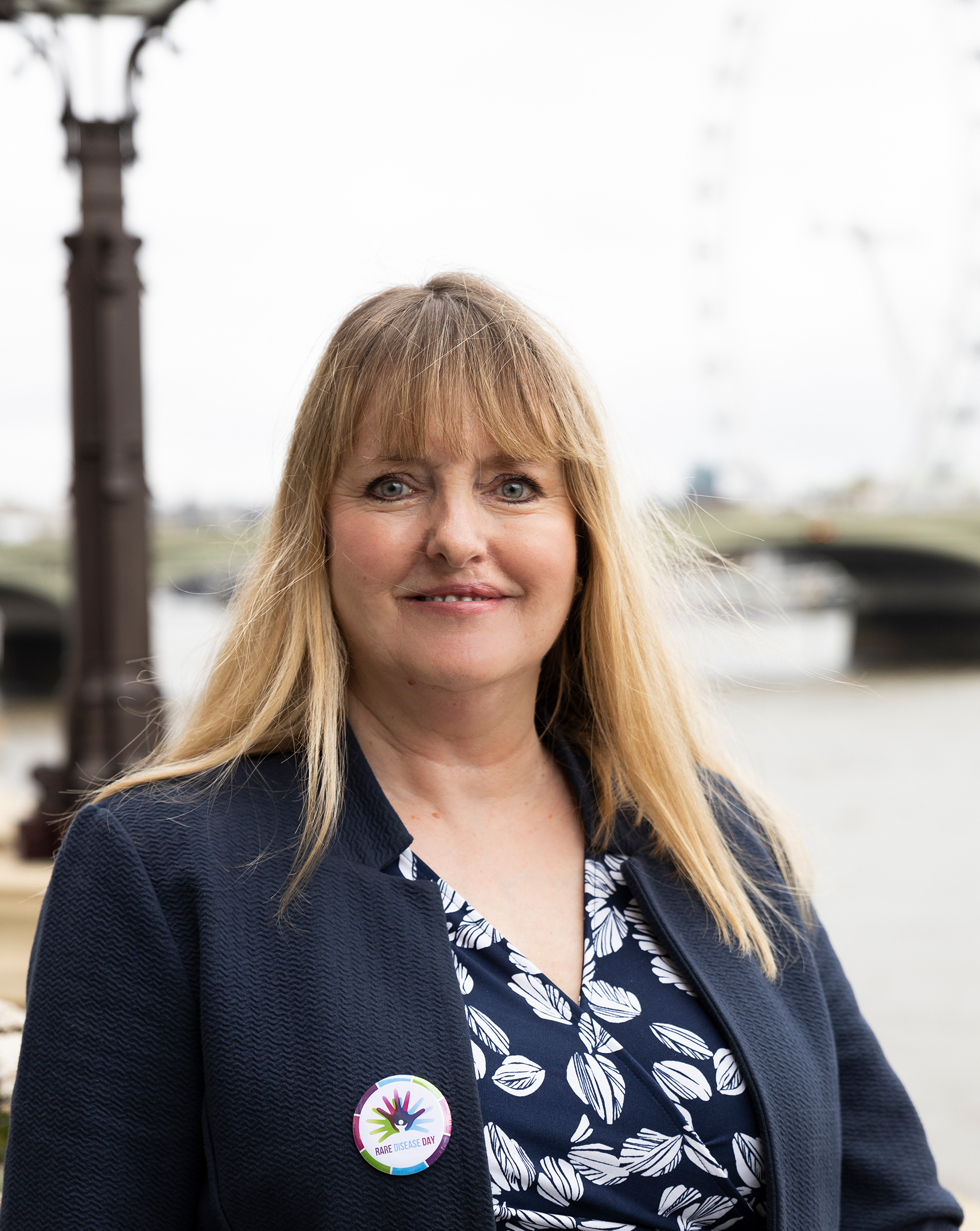 A headshot of Mary, Genetic Alliance UK's HR team. She has long blonde hair and wears a dark suit jacket and floral shirt. She stands by the Thames river at the Westminster Parliament building.