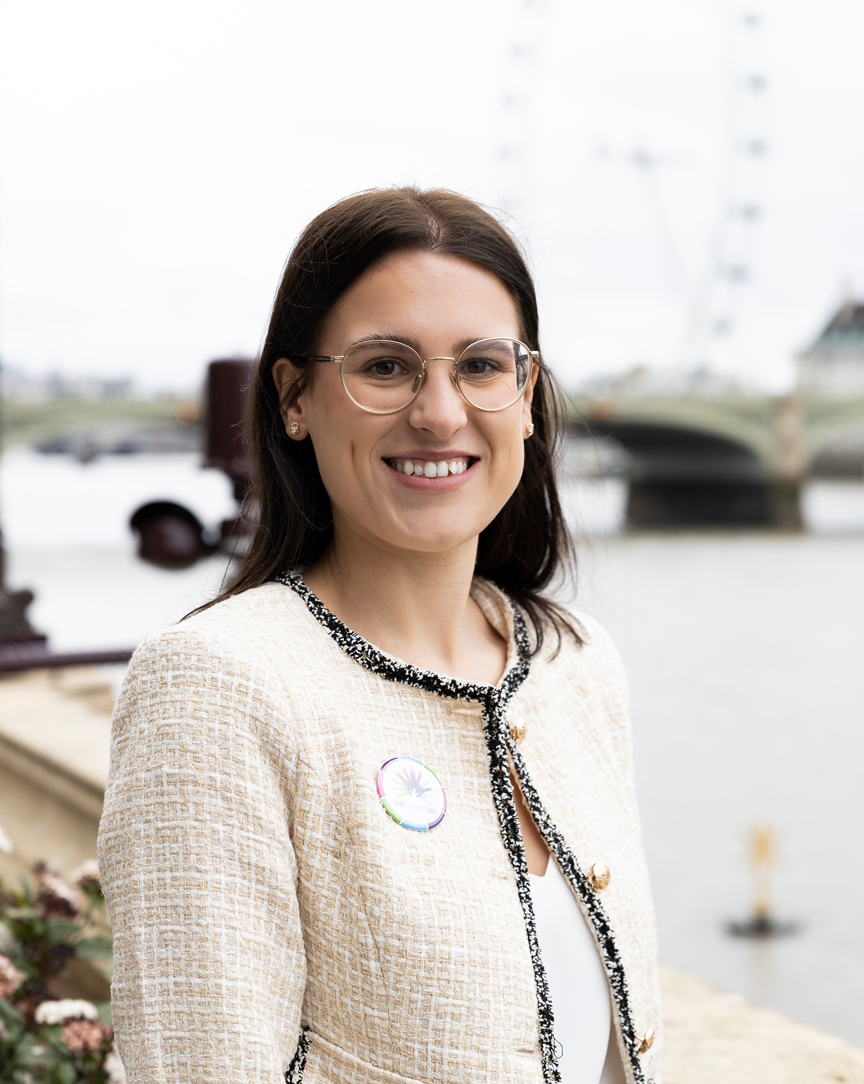 A headshot of Rachel, Genetic Alliance UK's Policy team. She has long brown hair that falls behind her shoulder. She wears a white cardigan and glasses. She stands by the Thames river at the Westminster Parliament building.