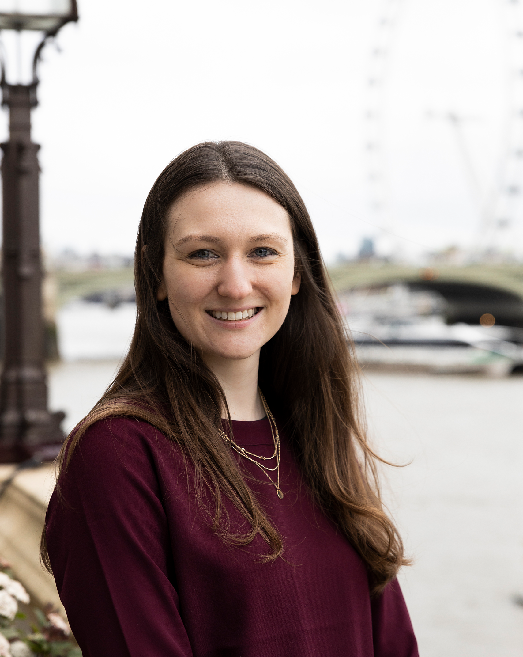 A headshot of Sophie, Genetic Alliance UK's Policy team. She has long brown hair and smiles with a friendly smile. She stands by the Thames river at the Westminster Parliament building.