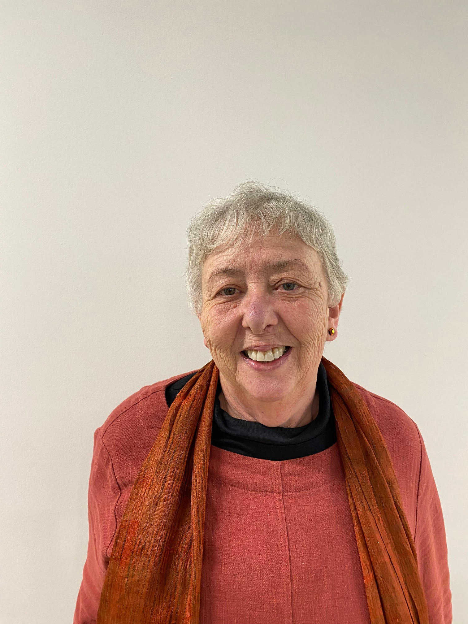 Sue Millman, an older woman with short grey hair. She wears a dark orange scarf and matching coloured jumper. She stands in front of a white wal