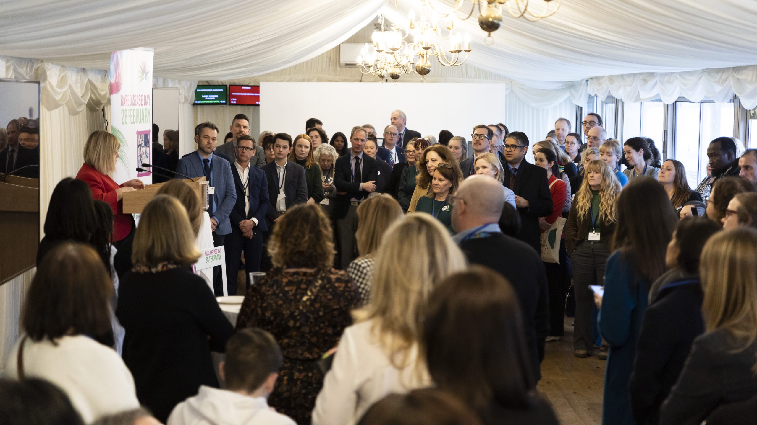 A collection of people around at the Genetic Alliance UK Rare Disease Day event in Westminster parliament. Everyone stands listening to a speaker, stood on a podium, to the left of the image