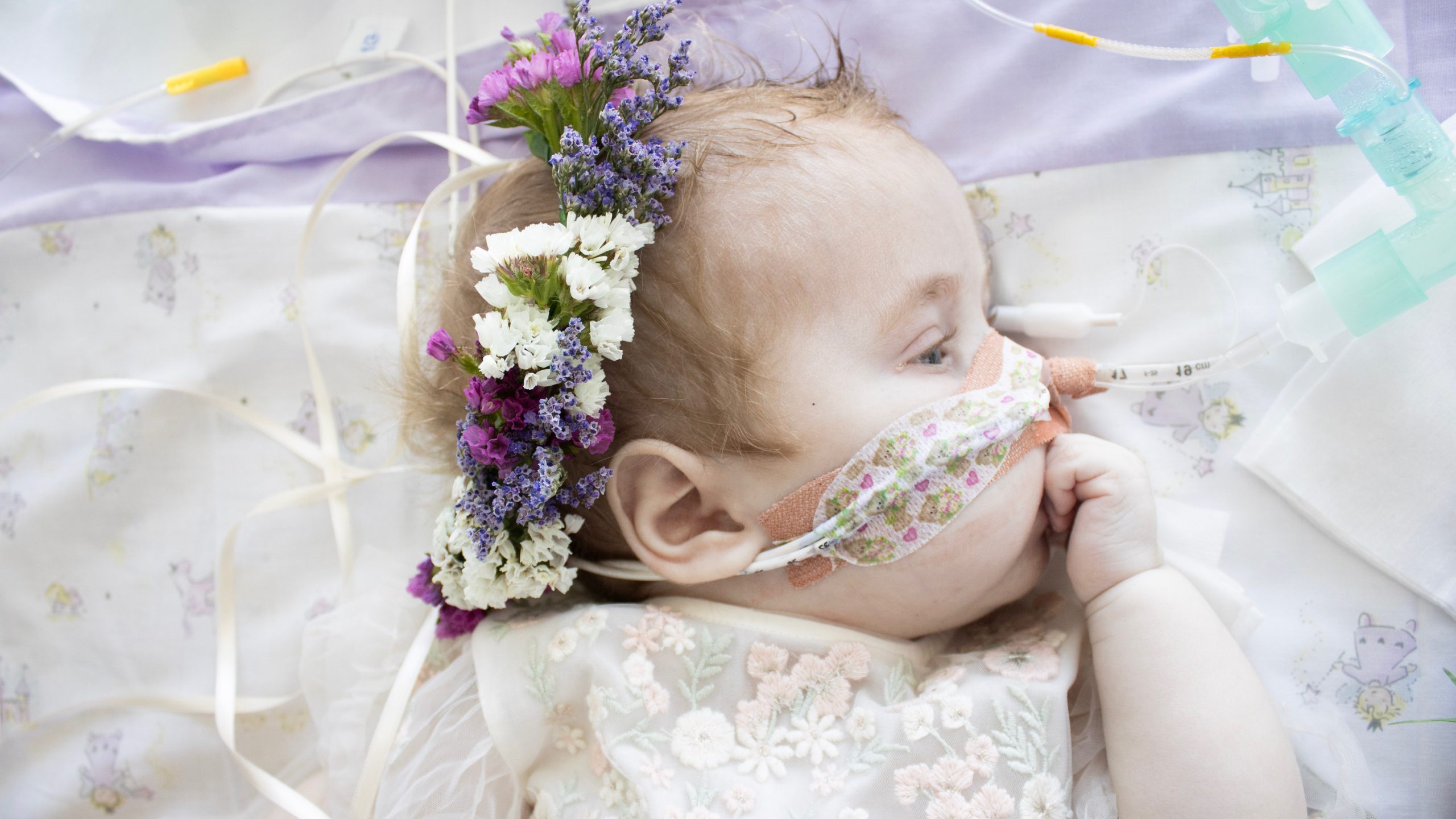 An image of a young baby in a bed. She wears a crown of purple flowers and looks to the side. Her hand is up to her mouth. She has a breathing tube taped to her face. The lilac bed sheets match the flowers in her hair.