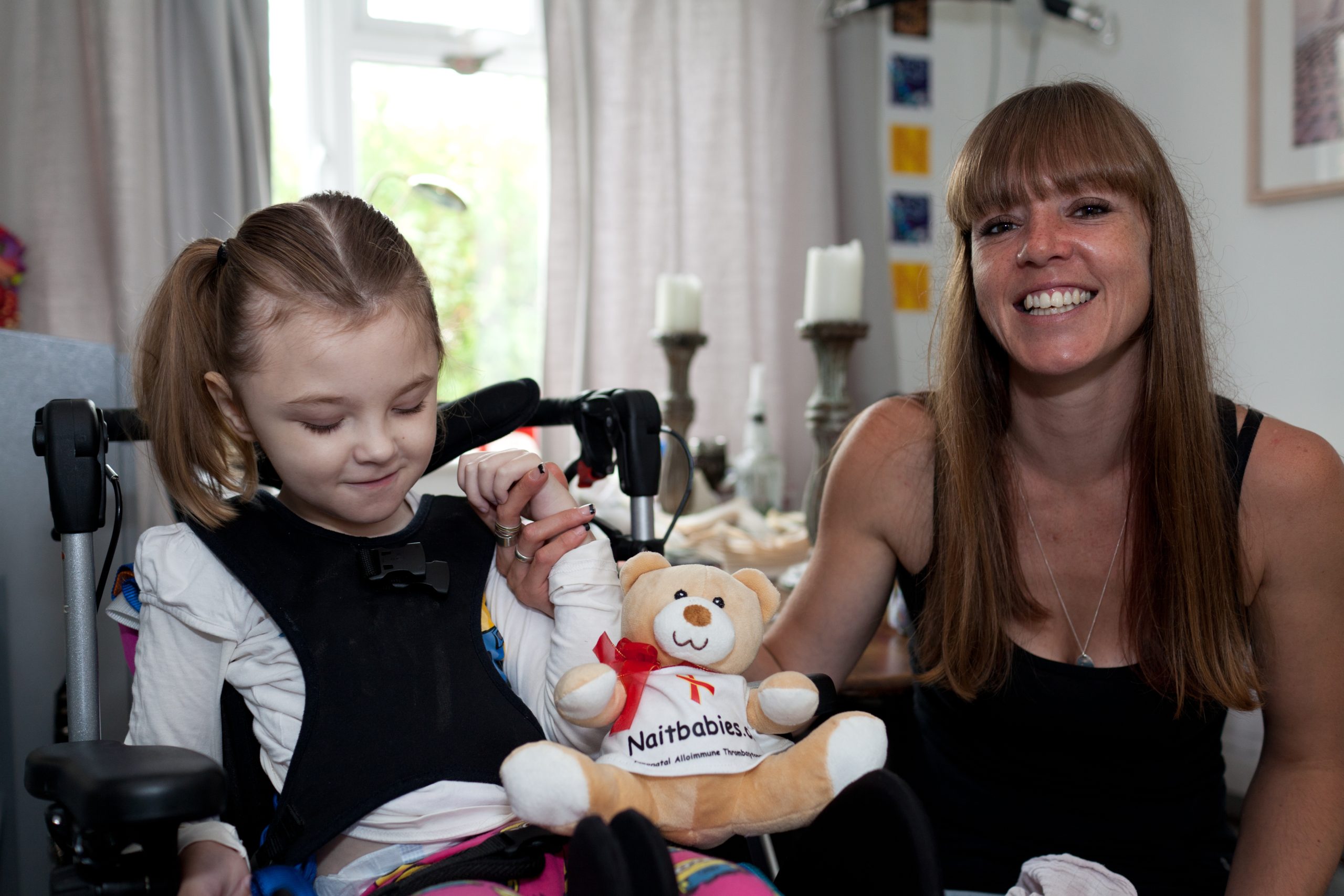 A girl sat in a wheelchair hold on to the hand of an adult woman with long straight hair. Between them sits a small toy bear that has 'naitbabies' written on it's small shirt.