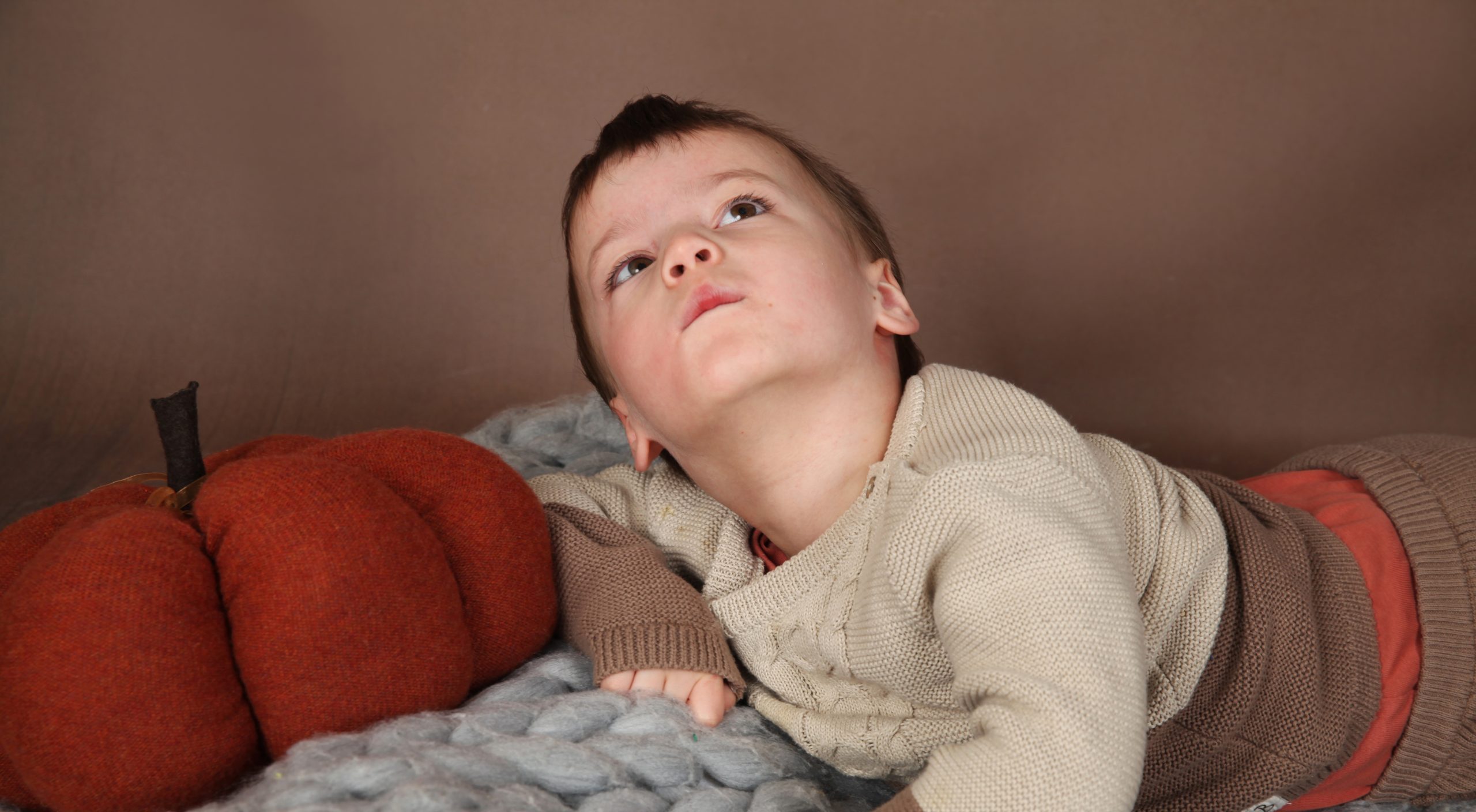 A young boy lying on is front. He props his head up and looks up to the sky looking to be in deep thought. A pumpkin made from material is sat next to him and he lies on a grey, knitted blanket.