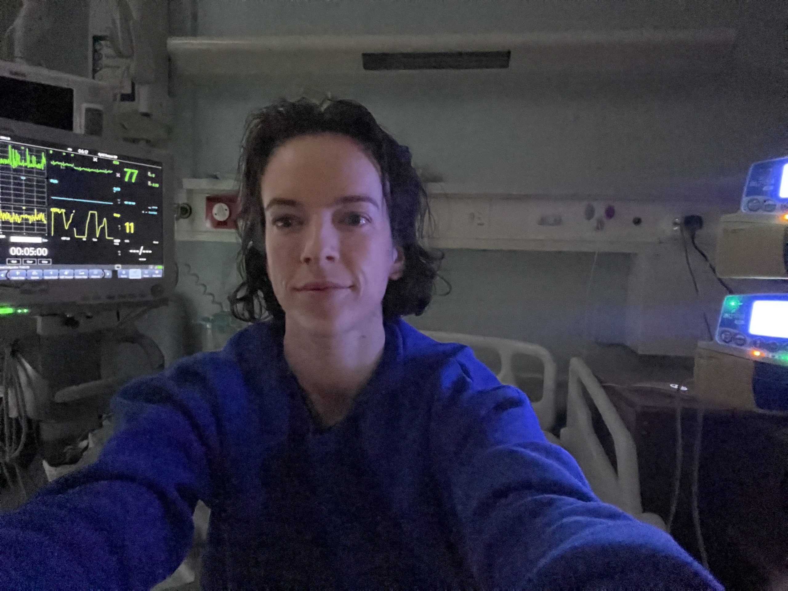 A photo of Dr Amber. She takes the photo of herself with her arms stretched out. She wears a blue, medical jacket in a hospital room