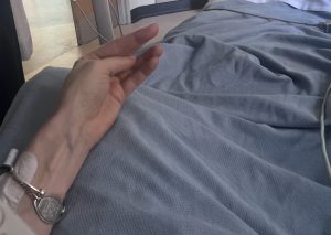A photo of a patient in a hospital bed, from their perspective. The photo looks down onto a cramped hand and blue hospital bed sheets