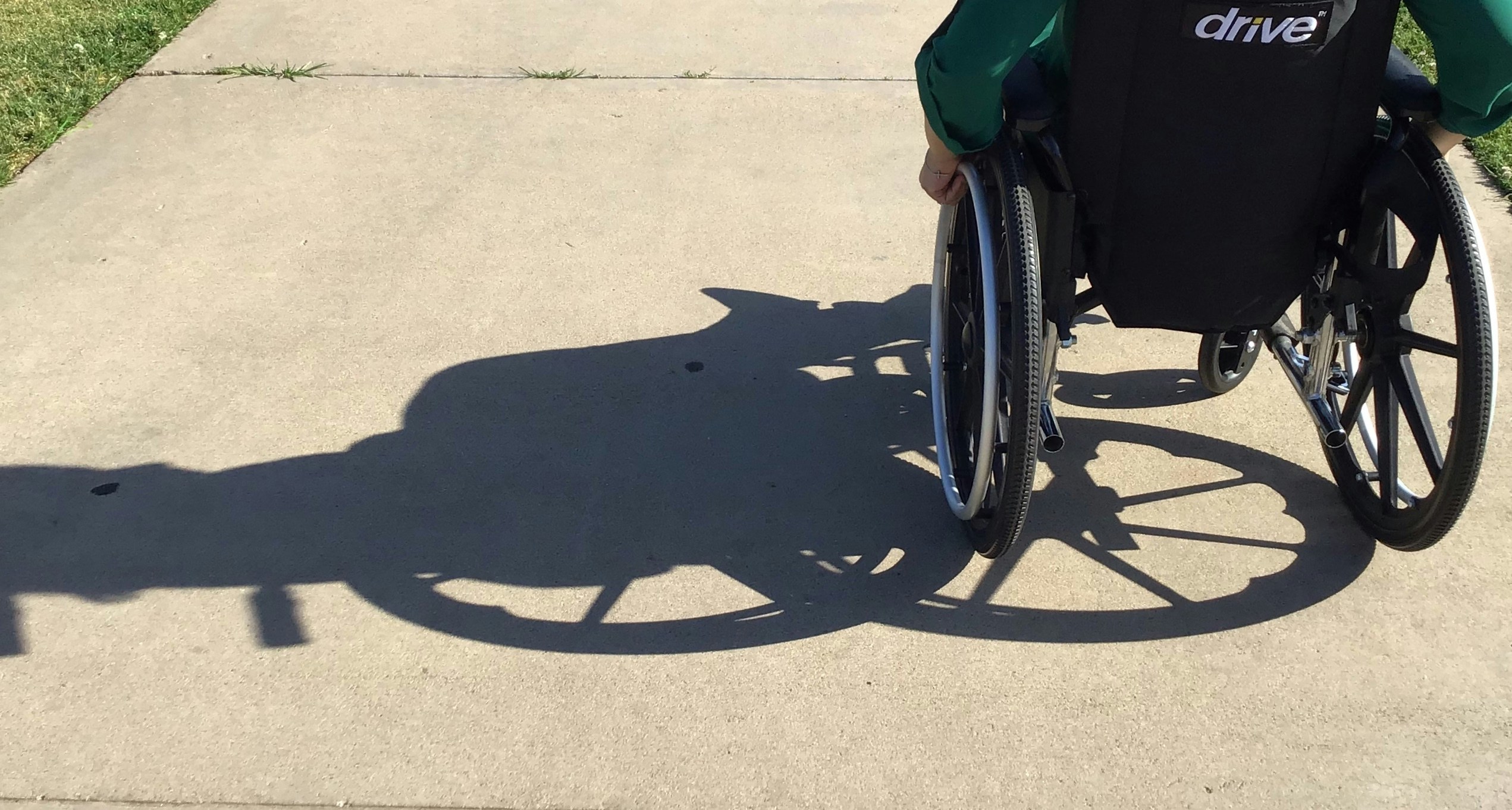 The bottom of a wheelchair with its shadow cast over the path it is being ridden on