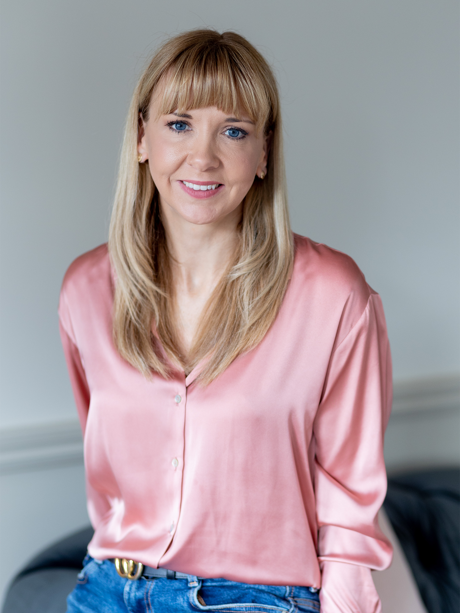 A woman in a silm light pink shirt and jeans. She has blonde hair and blue eyes and has a kind smile looking towards the camera